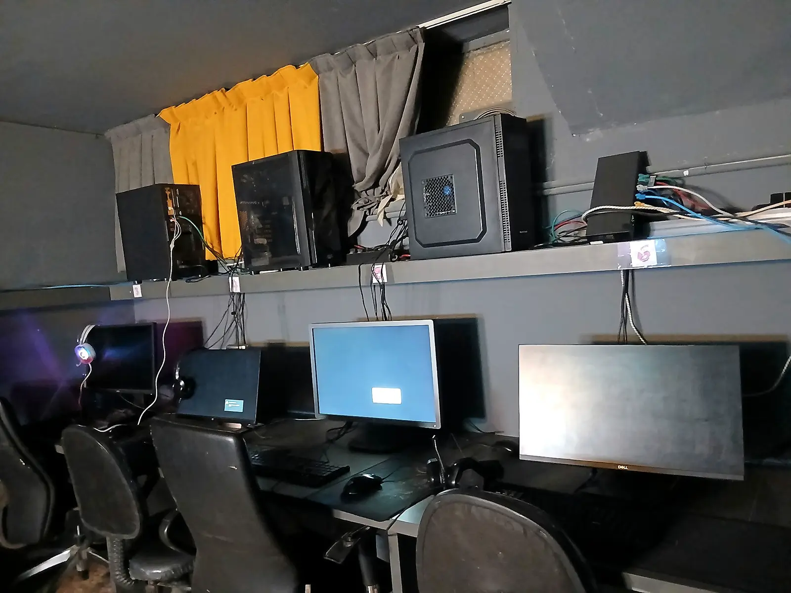 ردیف سیستم‌های PC گیم مسعود با کیس‌ها روی قفسه، نمایشگرها و صندلی‌های گیمینگ